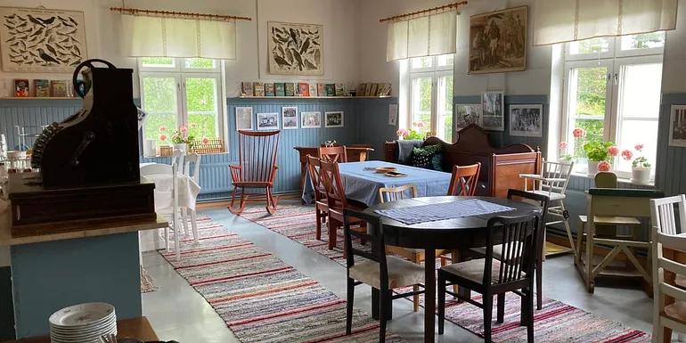 Kuva vanhasta luokkahuoneesta, joka sisustettu kahvilaksi vanhoilla huonekaluilla, pöytiä ja tuoleja, lattialla räsymatot.