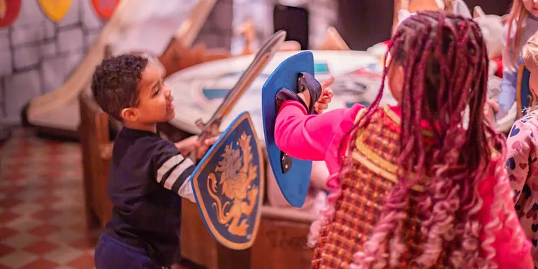 Two children are playfighting with soft swords in the room of King Arthur and his Knights.