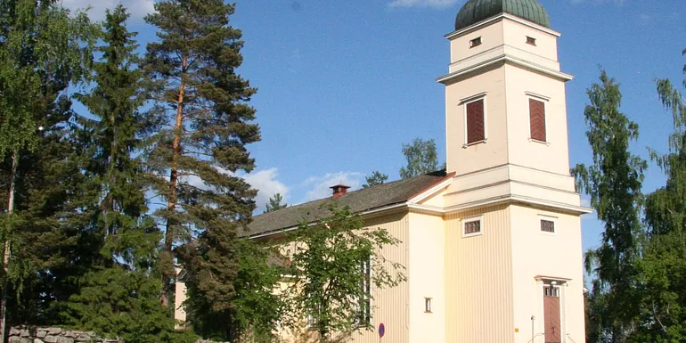 Viljakkalan kirkko kuvattuna kesällä ulkoapäin. Kirkko on kermanvaalea ja suoralinjainen rakennus, jonka kuvaa lähemmässä päädyssä kohoaa korkea torni.