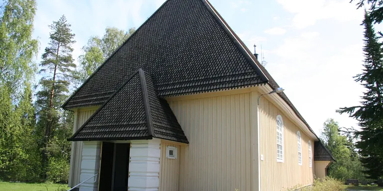 Aurejärven kirkko kuvattuna ulkoapäin. Rakennus on muodoiltaan pelkistetty, matalahko puurakennus, jossa on kermanvalkea verhoilu ja ruskeanharmaa viistokatto.
