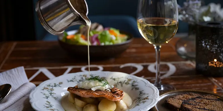 Hopea kannu, josta kaadetaan kastiketta kala-annoksen päälle. Anoksessa perunaa, fenkolia ja vaaleaa kastiketta. Taustalla valkoviinilasi ja toinen annos.