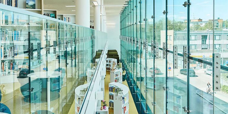 Kirjaston lasiseinän ja lasikaiteen välistä avautuu näkymä alakertaan lasten-ja nuorten lukutilaan.