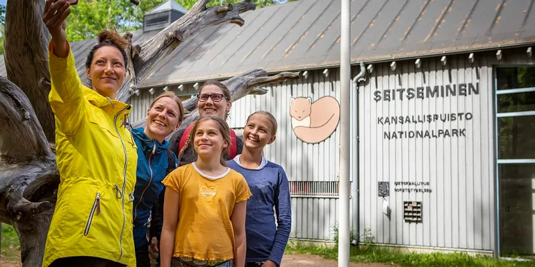 Kolme aikuista ja kaksi lasta ottamassa ryhmäkuvaa itsestään. Taustalla luontokeskuksen päärakennus. Seinässä lukee Seitseminen kansallispuisto.