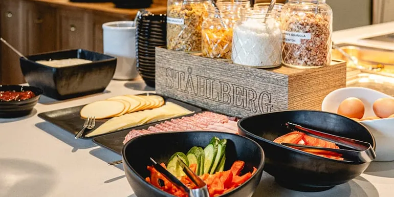 Aamiaisbuffet-tarjoilua pöydän päässä. Kuvan etualalla mustassa kulhossa kurkkua ja paprikaa, jonka takana leikkeleitä ja juustoja. Puisen korokkeen päällä granolaa ja siemeniä lasipurkeissa.