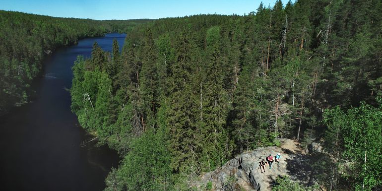 Ilmakuva kapeasta järvestä, jonka oikealla puolella kohoaa korkea kallio, muutoin järvi on metsien ympäröimä. Kalliolla istuu kolme retkeilijää. Kuva: Jari Salonen.