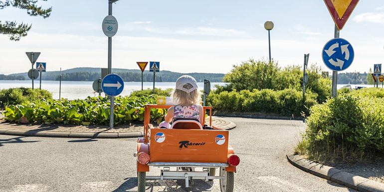 Lippalakkipäinen tyttö ajaa oranssilla polkuautolla liikenneympyrään. Taustalla järvi.